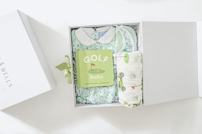 Decorative image open gift box with golf-themed baby dress, bloomers, and matching bib, golf-print swaddle blanket and "Golf Baby" board book. 