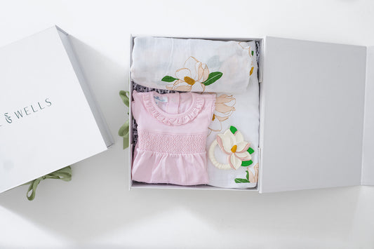 Open gift box with pink smocked bubble romper with a floral design, a magnolia-print muslin swaddle blanket, a matching burp cloth, and a magnolia-shaped baby teether.