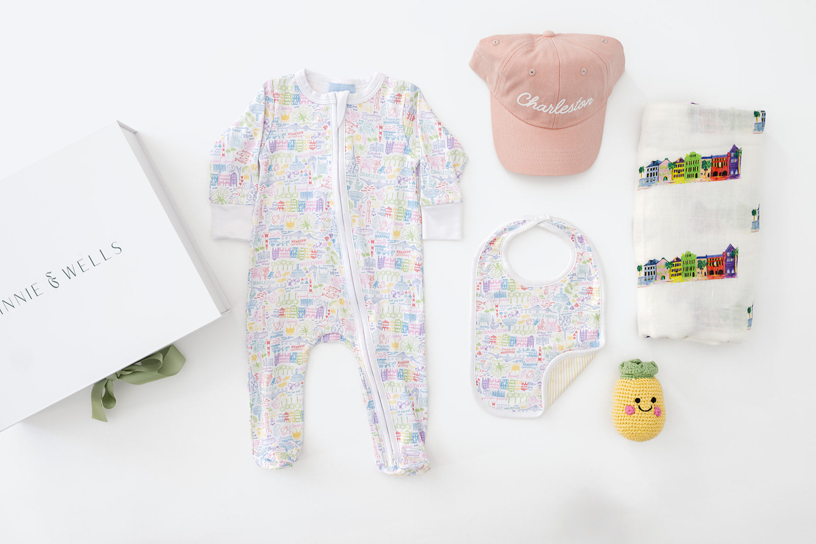 Baby clothing and accessories including a Charleston, South Carolina themed onesie, sleeper, hat, bib, and plush toy on a white surface.