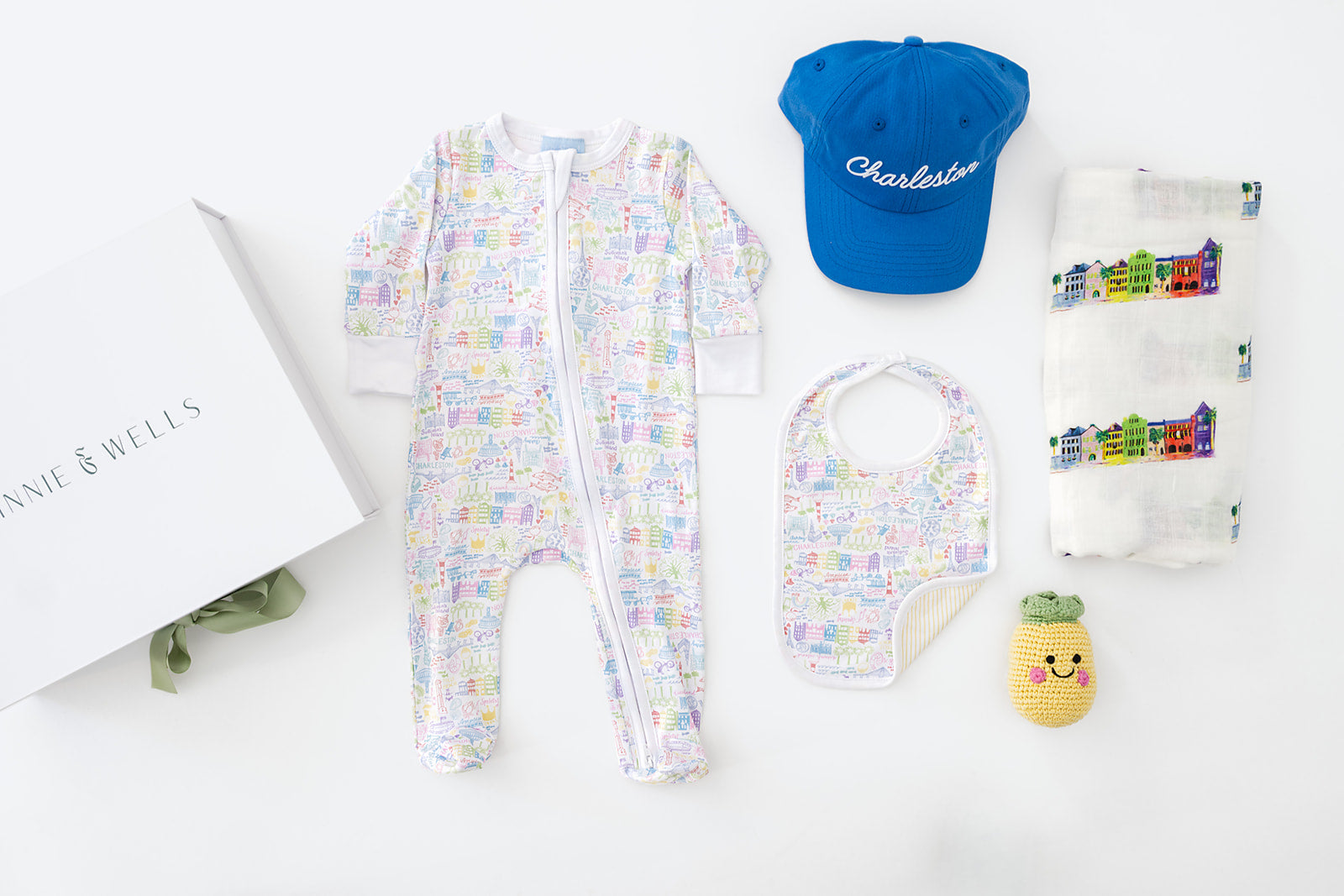 Baby clothing and accessories including a Charleston, South Carolina themed onesie, sleeper, hat, bib, and plush toy on a white surface.