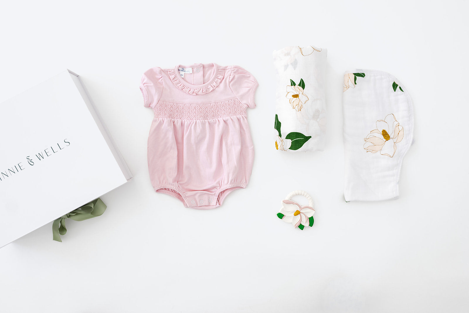 Decorative image lay out with a pink smocked bubble romper with a floral design, a magnolia-print muslin swaddle blanket, a matching burp cloth, and a magnolia-shaped baby teether.